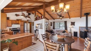 A rustic cabin interior with a wooden ceiling, a central dining area with a round table, a kitchen with wooden cabinets, and a fireplace. - No longer available for short term rental due to county restrictions (Captain Cook)