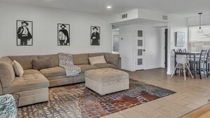 A modern living room with a sectional sofa, a matching ottoman, and a patterned rug. There is a dining area with a table and chairs visible in the background. - Urban Desert Home (Phoenix)