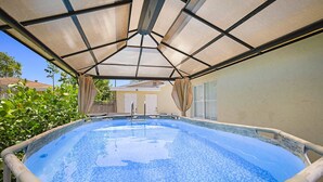 Une piscine extérieure surmontée d’une pergola, entourée de verdure et avec une maison en arrière-plan.