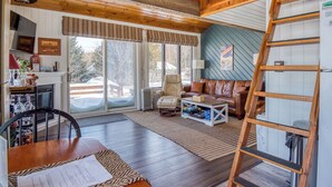 A cozy cabin interior with a dining area, a living room with a sofa and a coffee table, and a large window with blinds. - Crisp Cabin: Waterfront, 2 Kayaks, AC + Walk Downtown (Rangeley)