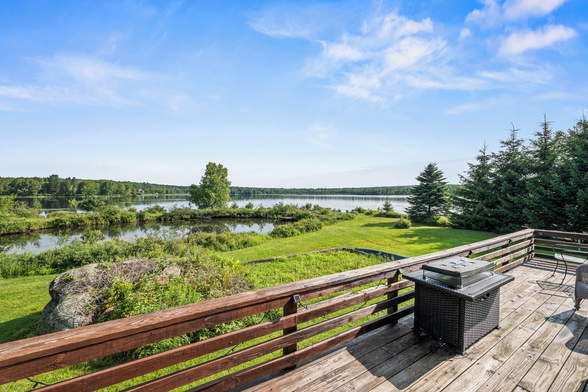 A wooden deck with a grill and seating area overlooking a lake with trees and a distant shoreline.