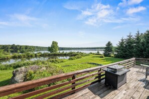 Une terrasse en bois avec un barbecue et un espace salon, donnant sur un lac bordé d’arbres et dont on aperçoit la rive au loin.