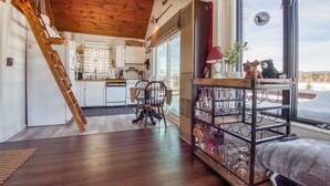 A cozy kitchen with wooden beams, a dining area with a chair, and a wine rack by the window. - Crisp Cabin: Waterfront, 2 Kayaks, AC + Walk Downtown (Rangeley)