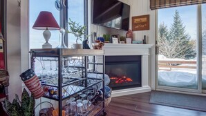 A cozy living room with a fireplace, a TV mounted above it, and a large window offering a view of snow-covered trees. - Crisp Cabin: Waterfront, 2 Kayaks, AC + Walk Downtown (Rangeley)