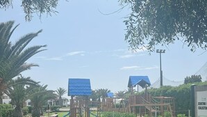 Un parque infantil con estructuras para escalar y toboganes, rodeado de césped cuidado y palmeras.