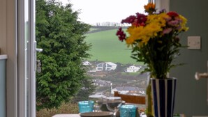 A dining area with a table set for two, a vase of flowers, and a view of houses and greenery outside. - Valley View by Interhome (Mevagissey)
