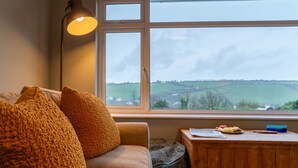 A cozy room with a large window, a sofa, a lamp, and a small wooden table with a view of the countryside. - Valley View by Interhome (Mevagissey)