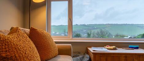 A cozy room with a large window, a sofa, a lamp, and a small wooden table with a view of the countryside.