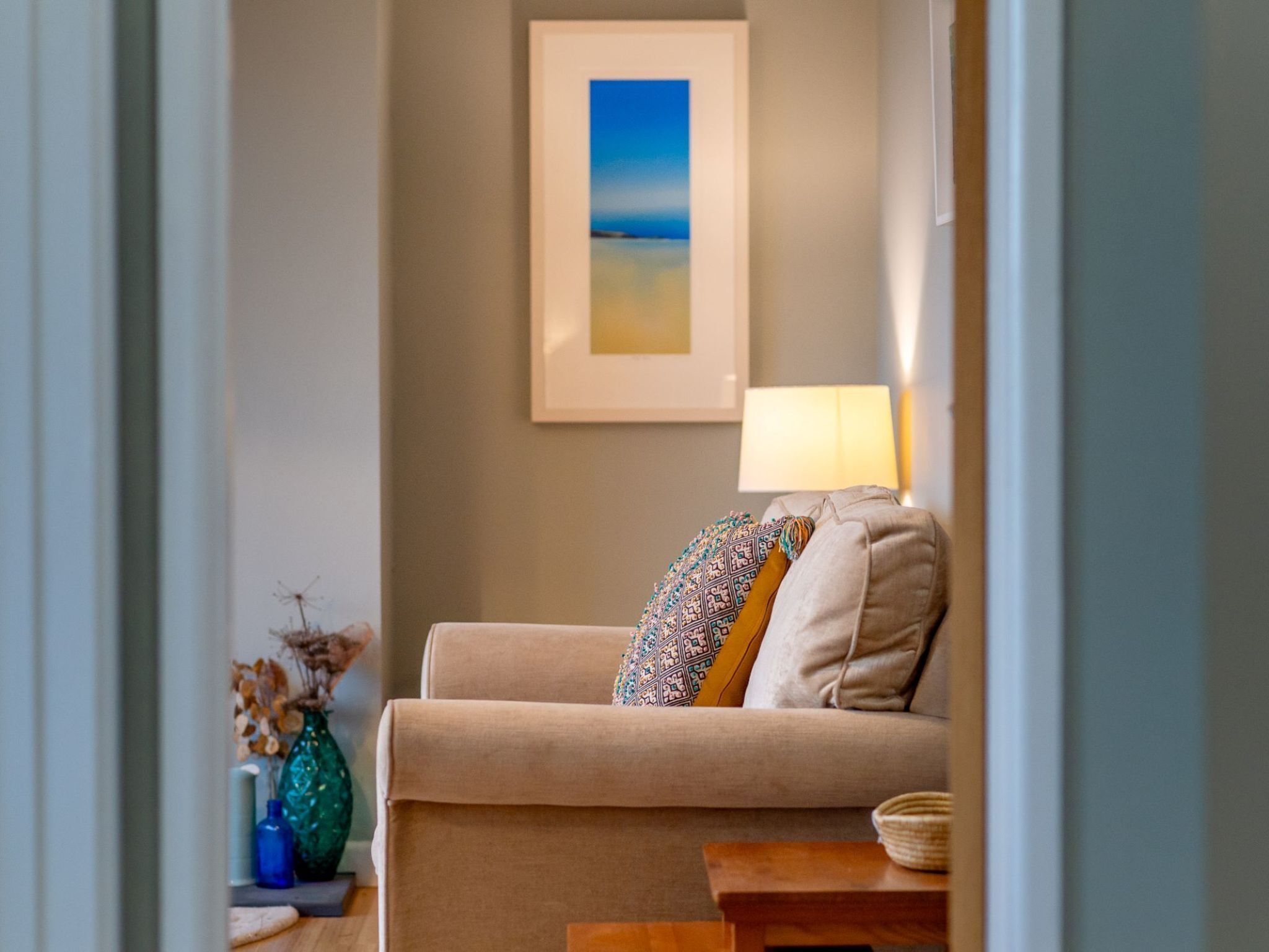 A cozy living room with a beige sofa, a patterned throw pillow, a framed painting of a beach, and a lamp on a side table.
