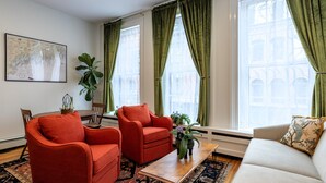 Una sala de estar con un sillón rojo, un sofá color beige y una mesa de centro. Hay dos ventanas con cortinas verdes, una planta grande y un mapa enmarcado en la pared.