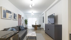 Una sala de estar moderna con sofá, mesa de centro y televisor. Hay un comedor con mesa y sillas.
