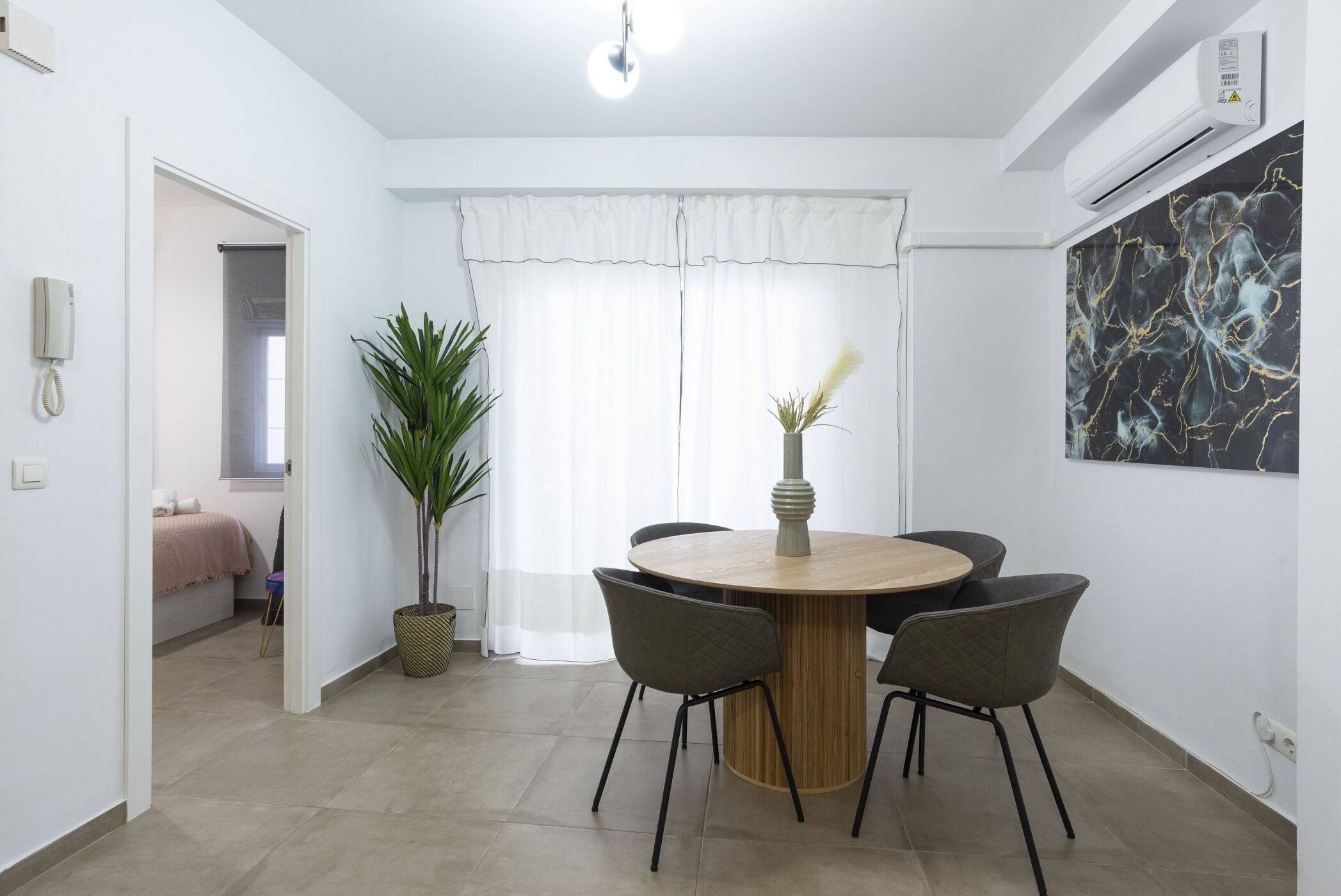A modern dining area with a round wooden table, four chairs, a potted plant, and a large abstract painting on the wall.