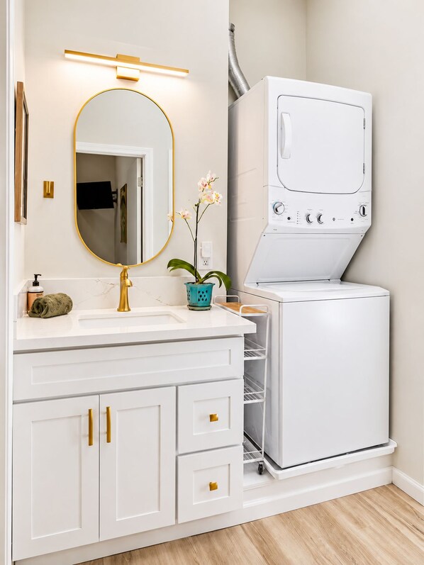 A bathroom with a white vanity, gold handles, a round mirror with a gold frame, a white washing machine, and a small orchid plant.