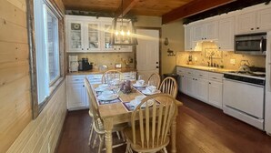 Une cuisine aux murs lambrissés, avec des meubles blancs, une table à manger dressée pour six personnes et une grande fenêtre sur le côté gauche.