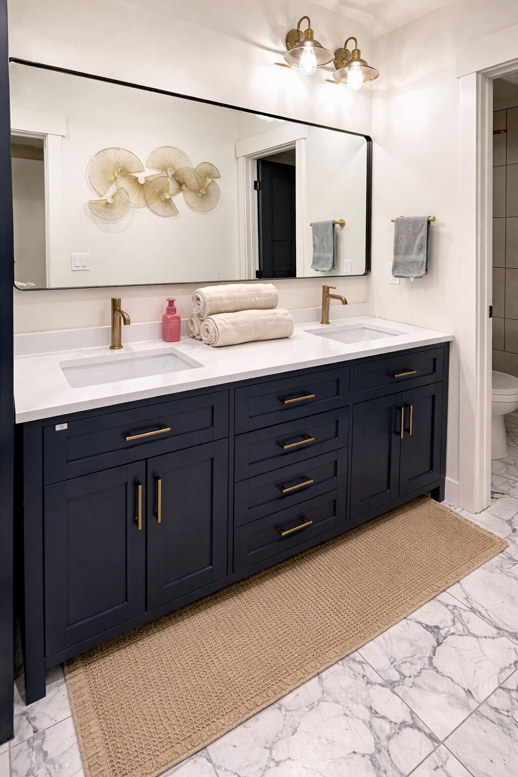 A bathroom with a double sink vanity, dark cabinets, gold handles, a large mirror, and wall-mounted fans.