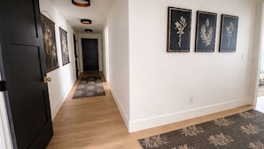 A hallway with a dark door, a light wooden floor, a dark rug, and three framed botanical prints on the wall. - Brand new apartment, sleeps 10, IDEAL location in Utah county. High end finishes (Lindon)