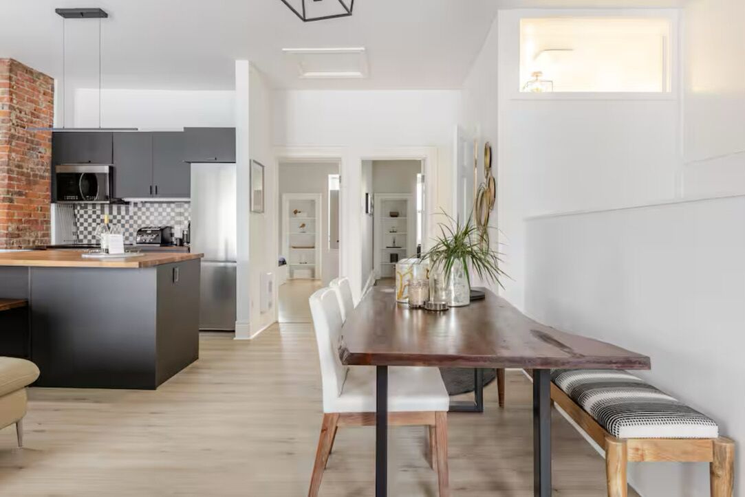 A modern kitchen with a central island, a dining area with a wooden table and benches, and a view of the adjacent living space.