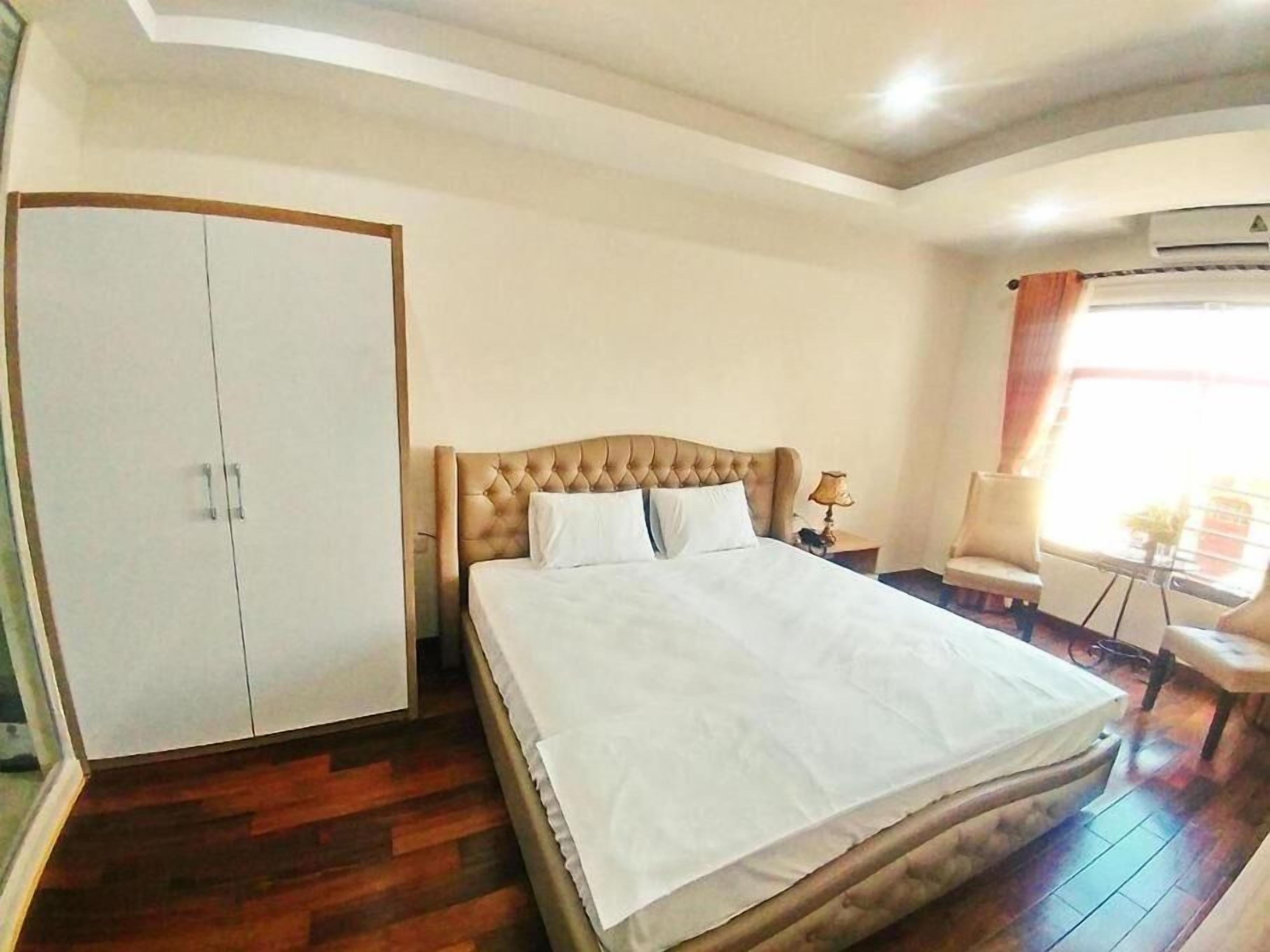 A hotel room with a large bed, a wardrobe, a small table with a lamp, and a chair.