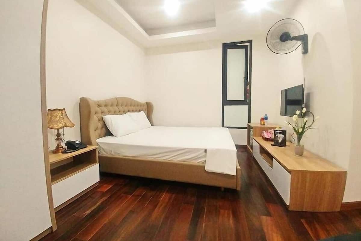 A bedroom with a bed, bedside table, lamp, dresser, mirror, and a fan.