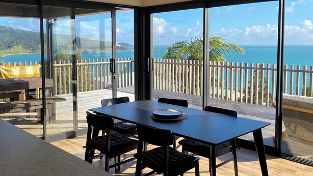 A modern dining area with a table and chairs, large windows offering a view of the sea and mountains, and a ceiling fan. - Ahipara Beach Pad (Ahipara)