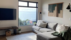 A modern living room with a large window, a grey sofa, a wooden floor, and a ceiling fan. - Ahipara Beach Pad (Ahipara)