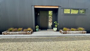 A modern building with a dark exterior, a wooden door, and two planters with plants on either side of the entrance. - Ahipara Beach Pad (Ahipara)