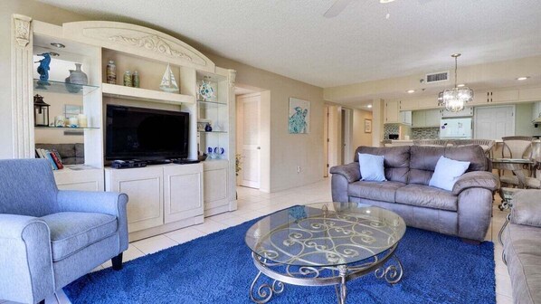 A living room with a TV, sofas, a glass coffee table, and a kitchen in the background. - First Floor, 10 Steps from the Pool & Ocean (New Smyrna Beach)
