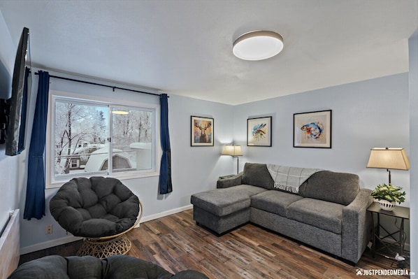 A modern living room with a grey sectional sofa, a round black chair, and a wooden floor. There are three framed artworks on the wall and a window with blue curtains.