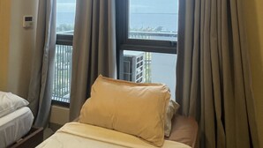 A hotel room with a bed, an air conditioning unit, a window with curtains, and a view of a body of water and buildings. - condominium rentals at Mactan Newtown (Lapu-Lapu)