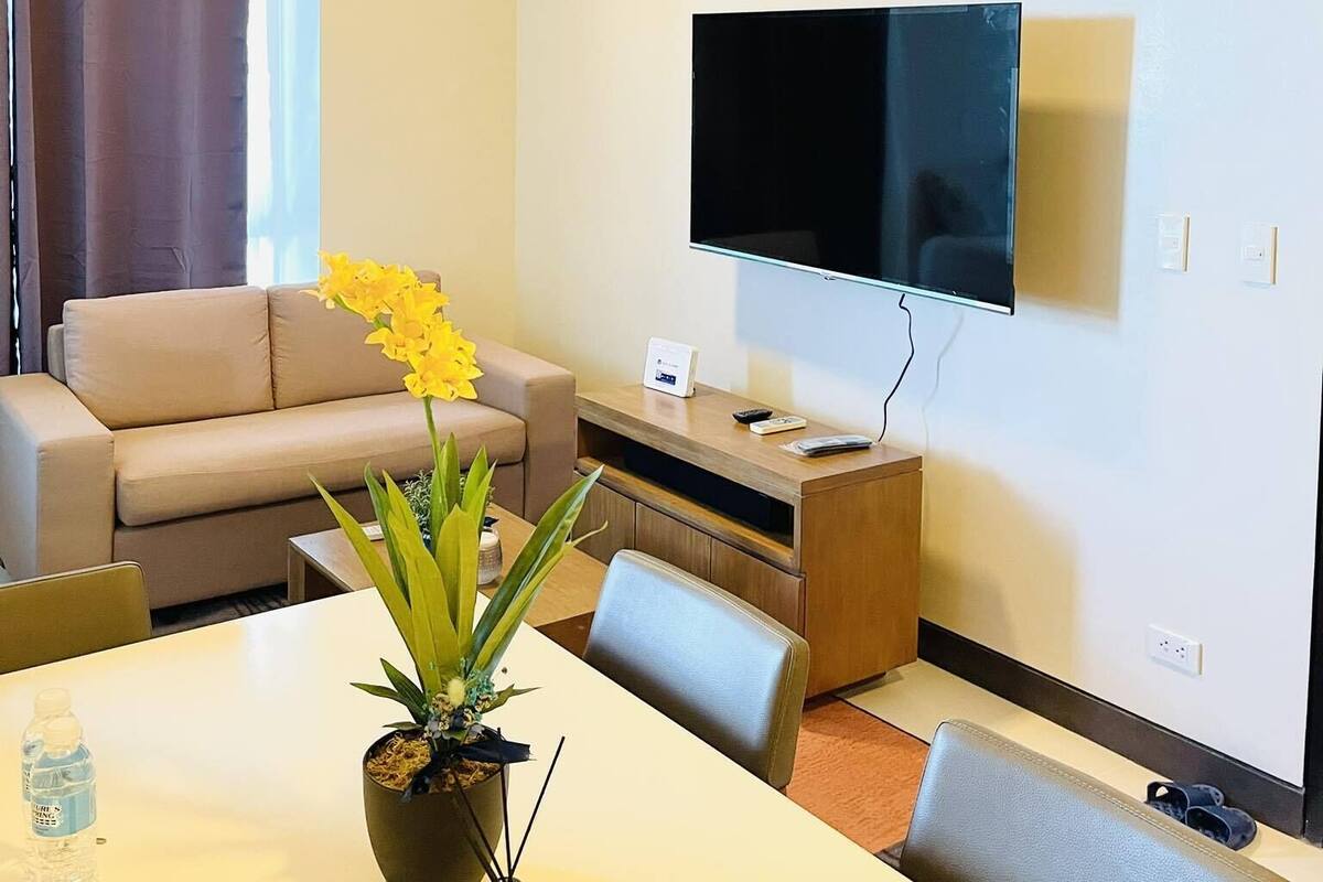A modern living room with a flat-screen TV, a sofa, a dining table with chairs, and a potted plant.