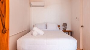 A neatly made bed with white linens, a ceiling fan, a small bedside table with a lamp, and an air conditioning unit on the wall. - Great location, walking distance to the beach -E (Puntarenas)