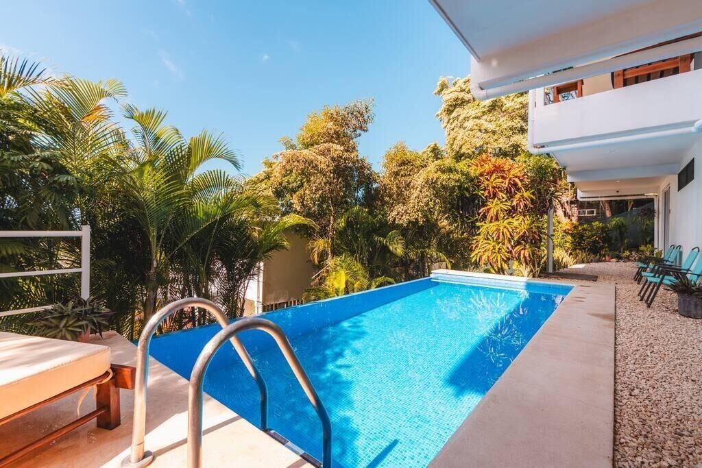 A rectangular swimming pool with a metal ladder, surrounded by tropical vegetation and a white building with a balcony.