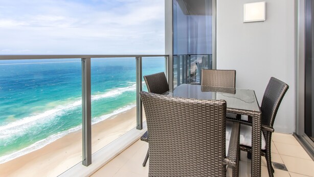 A balcony with a glass table and wicker chairs overlooking a beach and ocean.