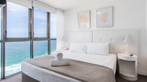 A hotel room with a large window overlooking the ocean, a bed with white linens, a bedside table with a lamp, and two framed pictures on the wall.