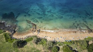 Vue aérienne d’une plage aux eaux cristallines, bordée d’un rivage sablonneux et parsemée de parasols.