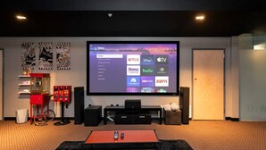 Un salón de cine en casa con un televisor de pantalla plana grande mostrando la interfaz de Roku, un mueble rojo con puerta de vidrio y un equipo de entretenimiento negro.