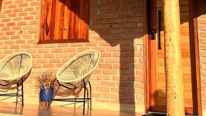 A rustic brick building with a wooden porch, two wicker chairs, and a potted plant. - Pousada Rota nas Montanhas (Camanducaia)