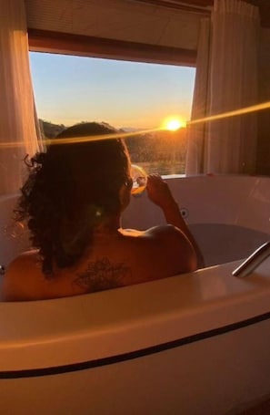 A person sitting in a bathtub with a view of the sunset through a window. - Pousada Rota nas Montanhas (Camanducaia)