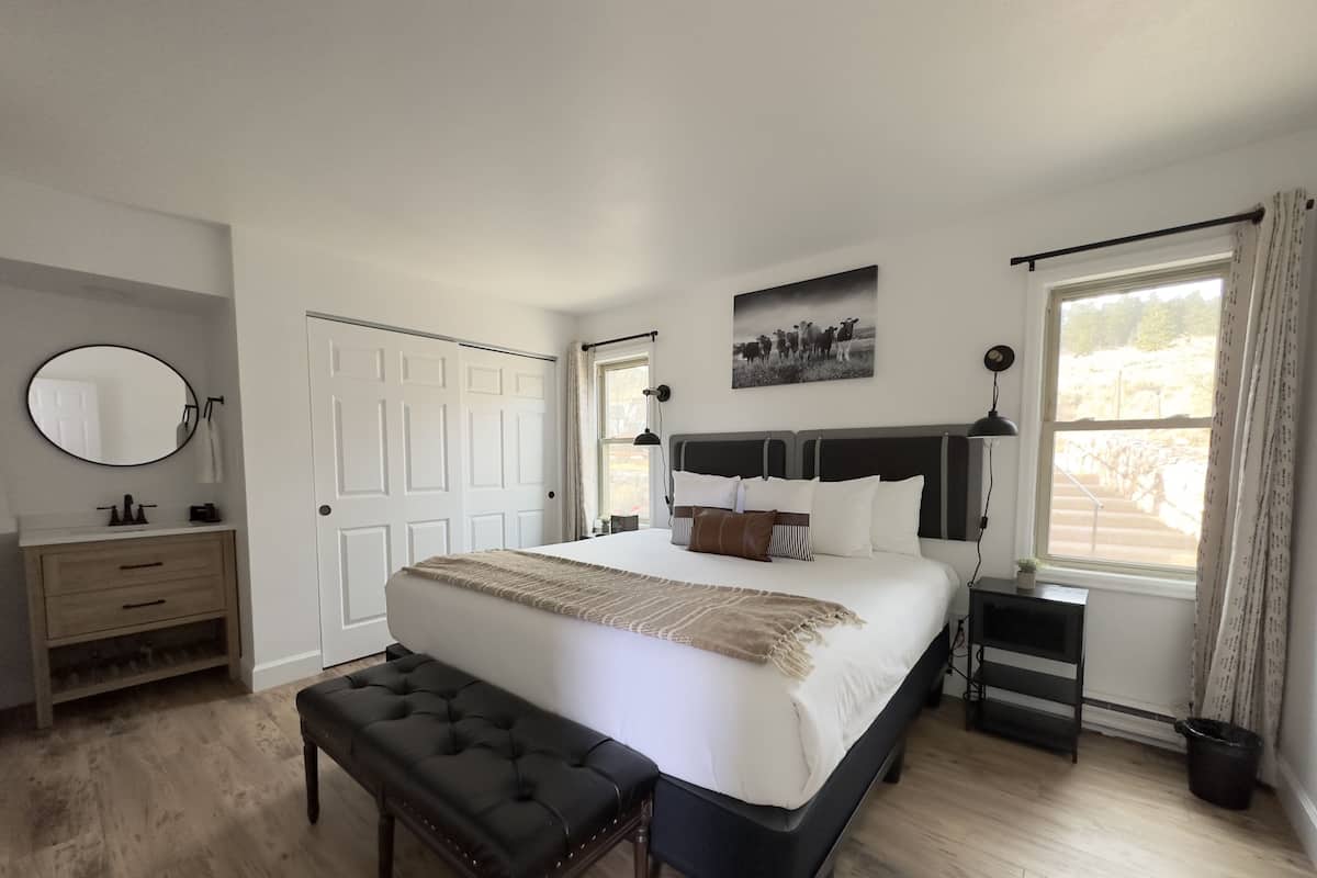 Un dormitorio con una cama grande, una mesita de noche, un banco, un aparador, un espejo y una ventana con cortinas.