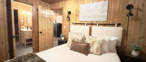 A bedroom with a wooden paneled wall, a bed with white bedding, a fluffy throw, and a painting above the bed.