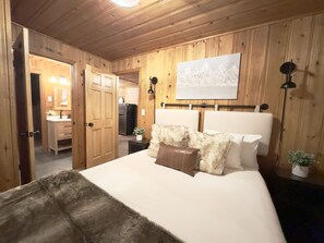 A bedroom with a wooden paneled wall, a bed with white bedding, a fluffy throw, and a painting above the bed.