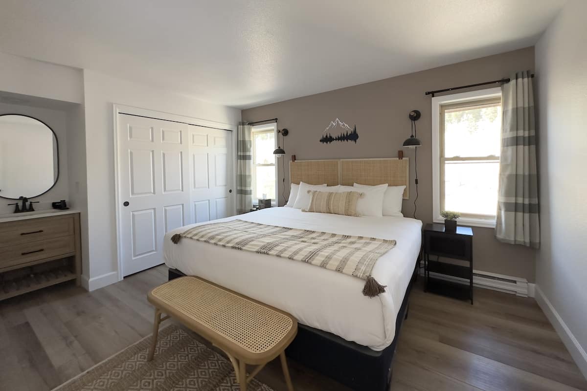 Un dormitorio con cama, mesitas de noche, un banco, cómoda, espejo y ventana con cortinas.