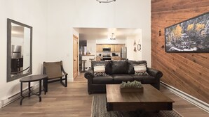 A modern living room with a leather sofa, wooden coffee table, and a dining table with chairs. A kitchen is visible in the background with wooden cabinets and a hanging light fixture. - Beaver Meadows Resort Ranch (Red Feather Lakes)