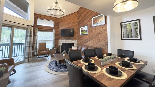 A modern dining area with a wooden table set for six, a fireplace, and a large window overlooking a deck. - Beaver Meadows Resort Ranch (Red Feather Lakes)