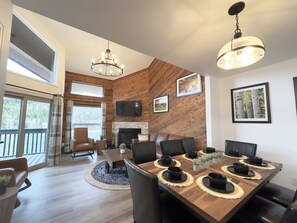 A modern dining area with a wooden table set for six, a fireplace, and a large window overlooking a deck. - Beaver Meadows Resort Ranch (Red Feather Lakes)