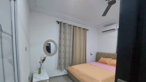 A bedroom with a bed, a bedside table with a vase, a mirror, and a window with curtains. - Monroe Luxury Apartments (Kumasi)