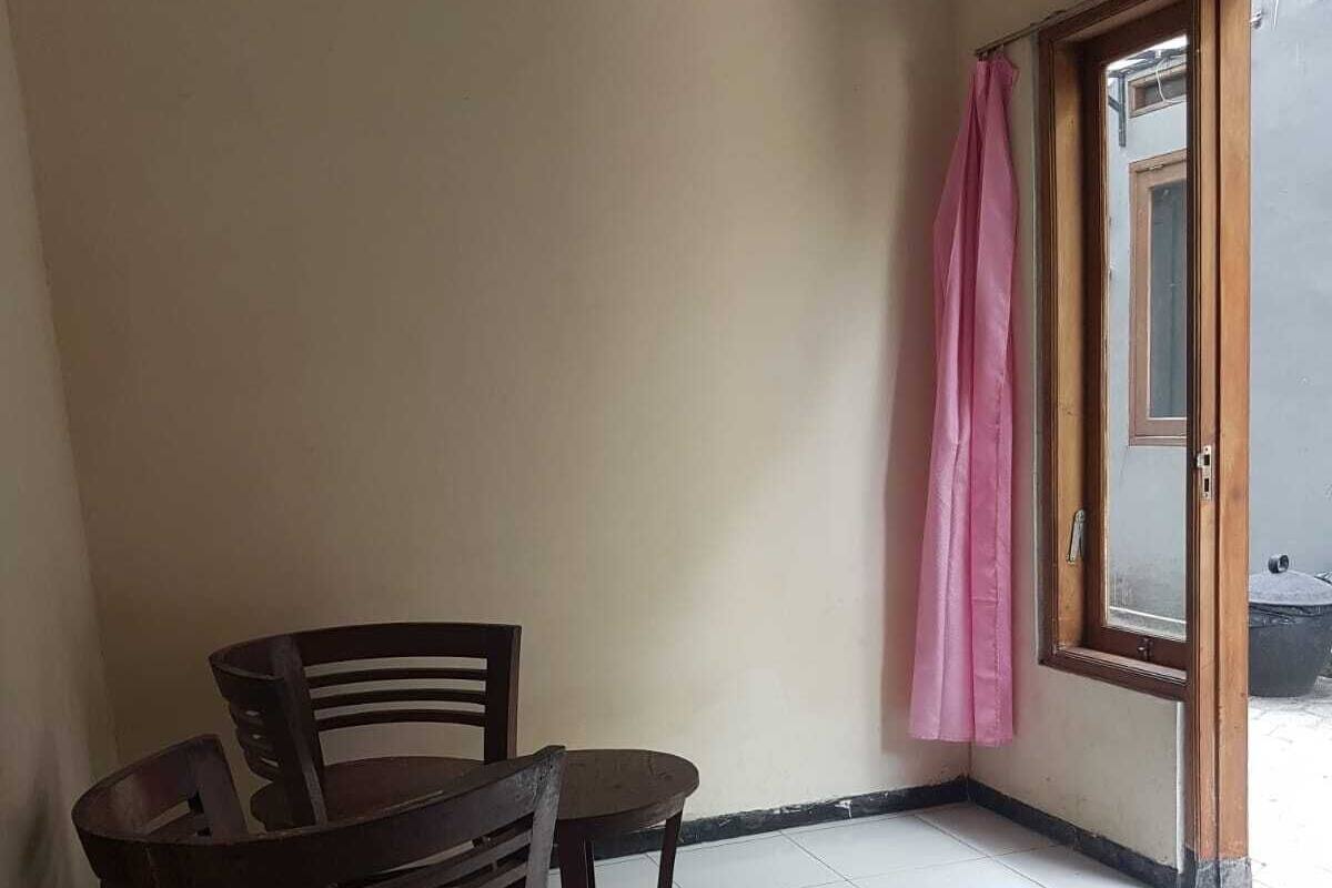 Una habitación con una silla de madera y una mesita pequeña, una puerta de vidrio corredera con una cortina rosa y piso de baldosas.