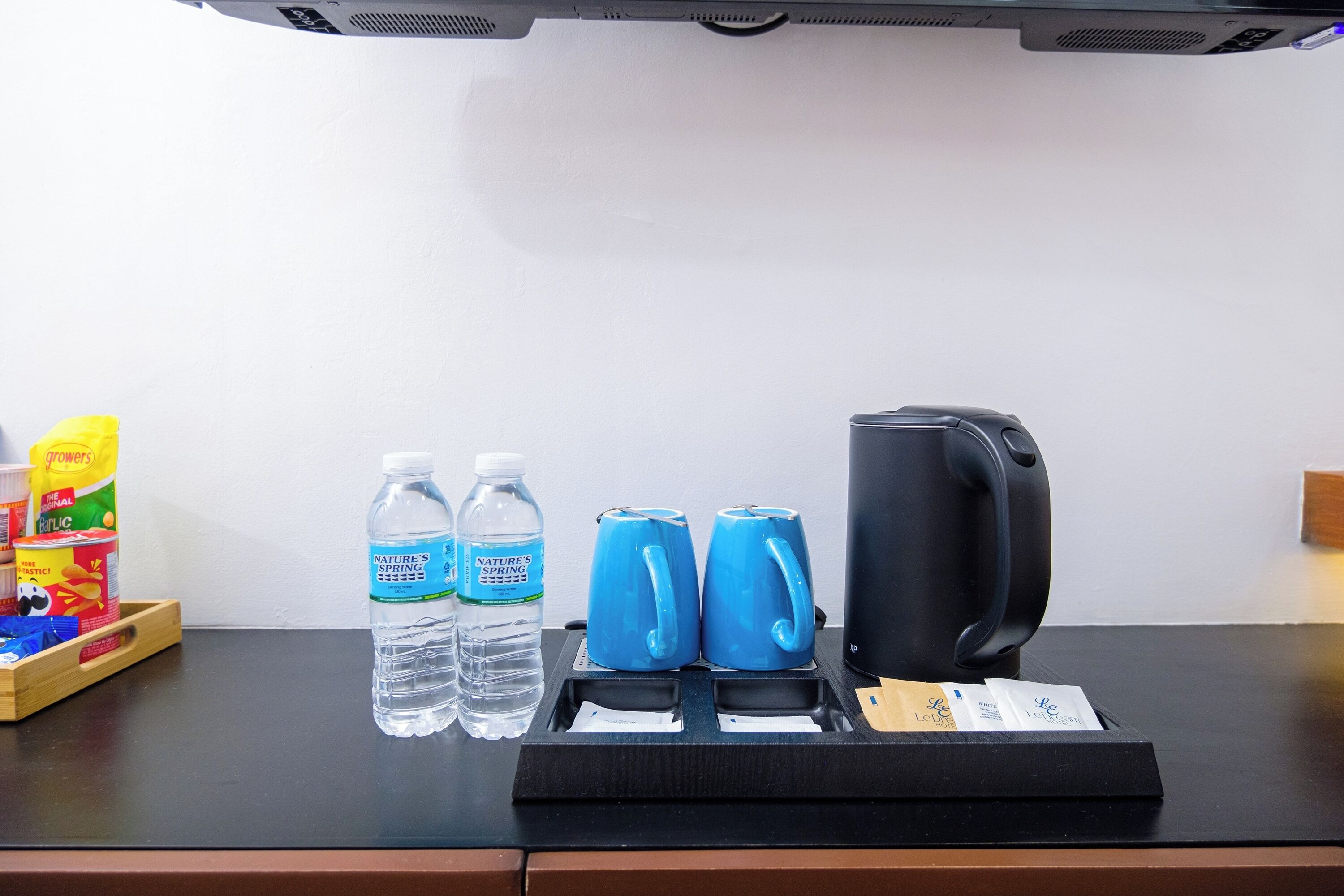 A hotel room's coffee station with two water bottles, two blue mugs, a black coffee maker, and a tray with two packets of tea.