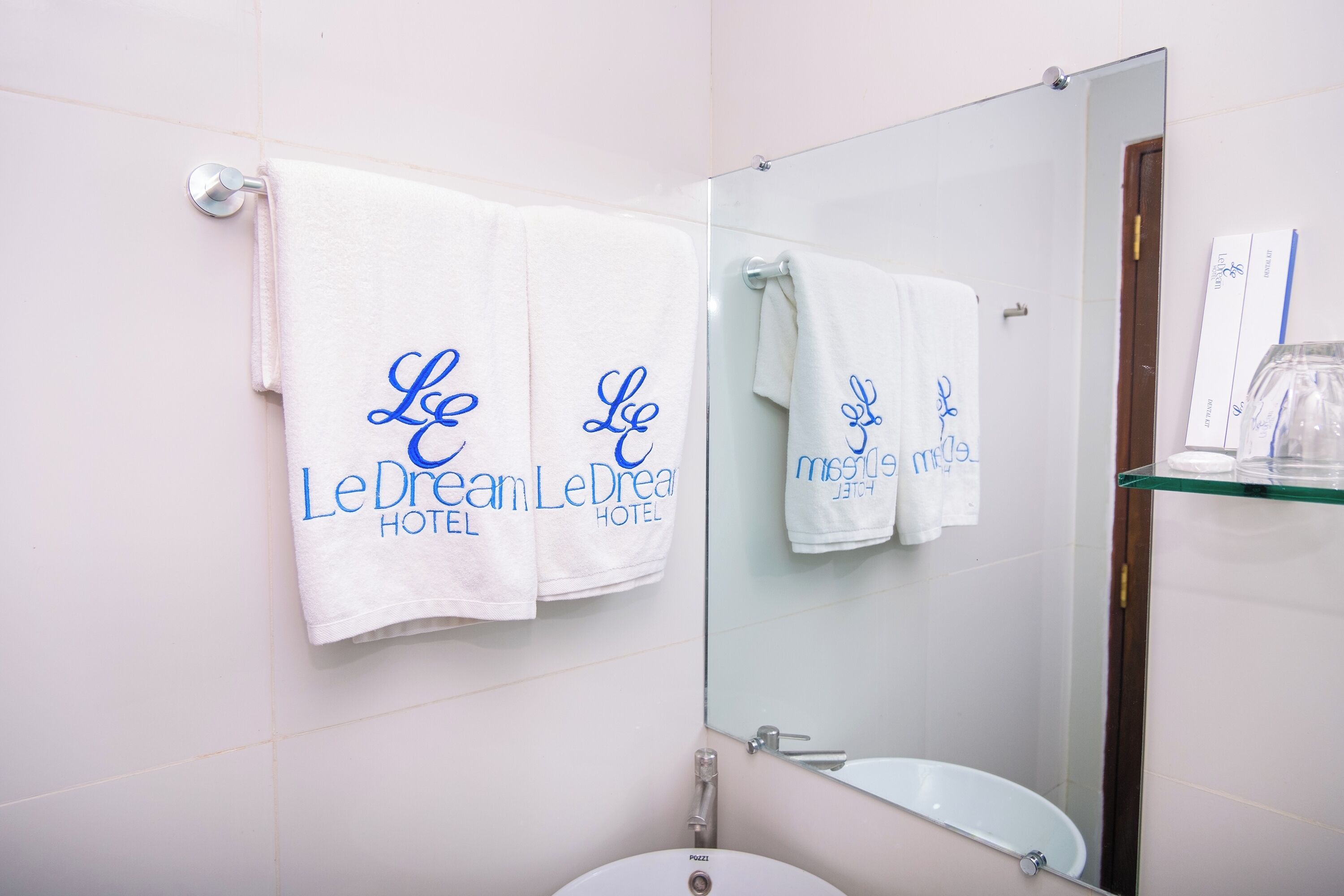 Une salle de bain carrelée de blanc, avec un lavabo, un miroir et deux serviettes accrochées au mur, sur lesquelles figure le nom de l’hôtel.