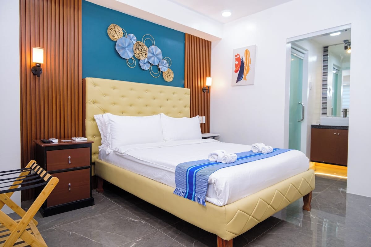 A modern hotel room with a large bed, wooden paneling, a decorative wall piece, and a small desk with a lamp.